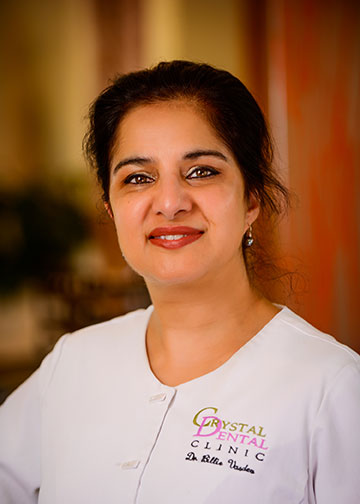 A woman posing professionally with a warm smile, wearing a white lab coat with name badge, set against a blurred indoor background.