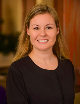 The image shows a woman smiling at the camera, wearing a dark top with a light-colored collar, standing against a blurred background that suggests an indoor setting with greenery and warm lighting.