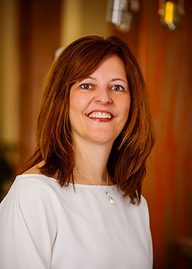 The image shows a woman with short brown hair, wearing a white top, standing against a light-colored wall with a framed picture above her head. She has a bright smile and appears to be posing for the photograph.