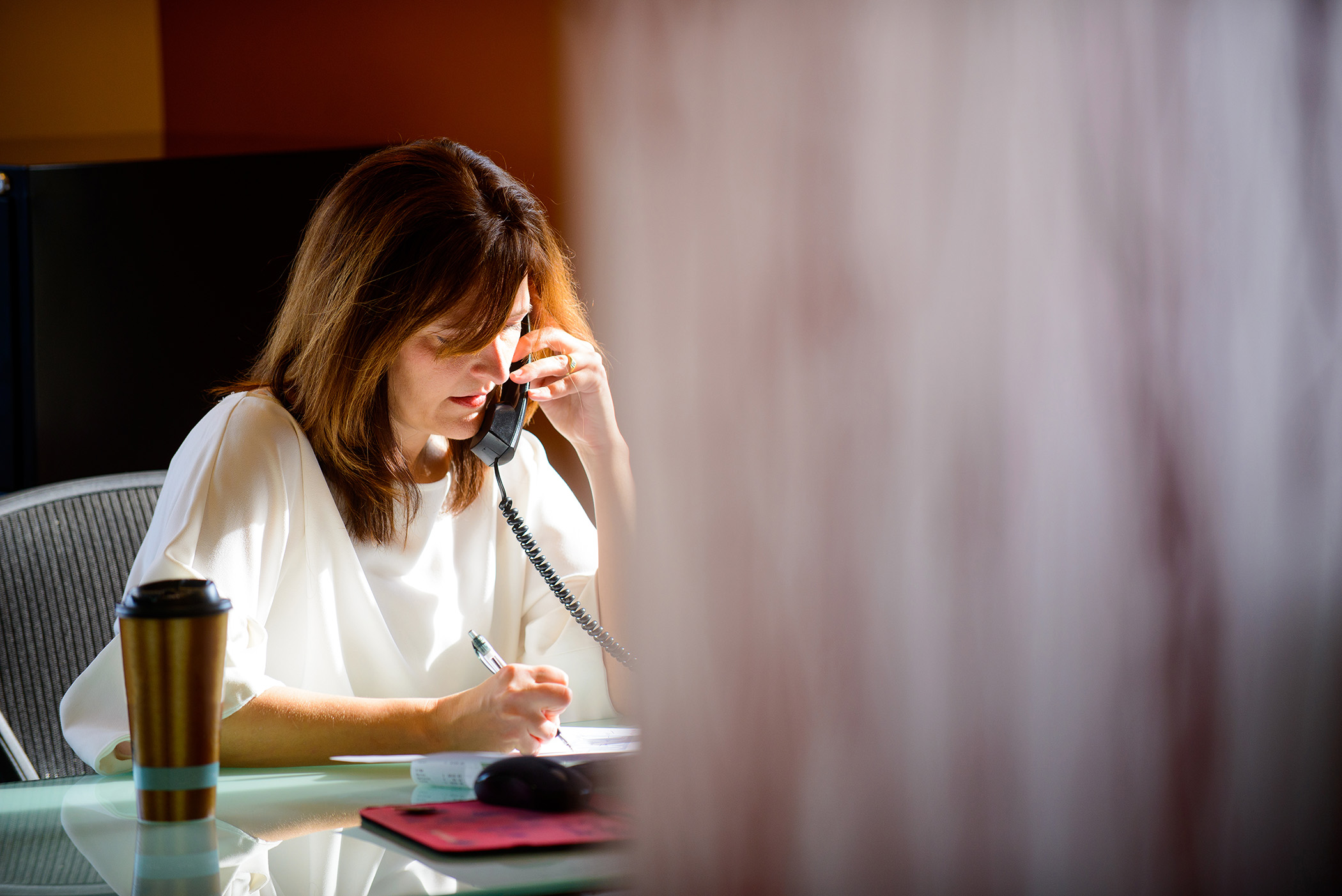 A woman sitting at a desk, talking on a phone while holding a pen, with a computer monitor and keyboard in front of her.