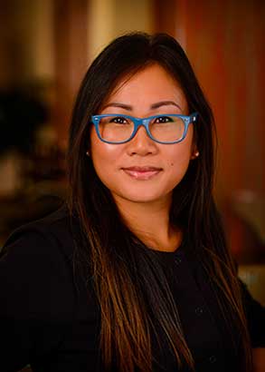 The image shows a woman with glasses posing for the camera, wearing a dark top and smiling slightly.