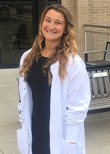 A person standing outdoors wearing a white lab coat and smiling at the camera.