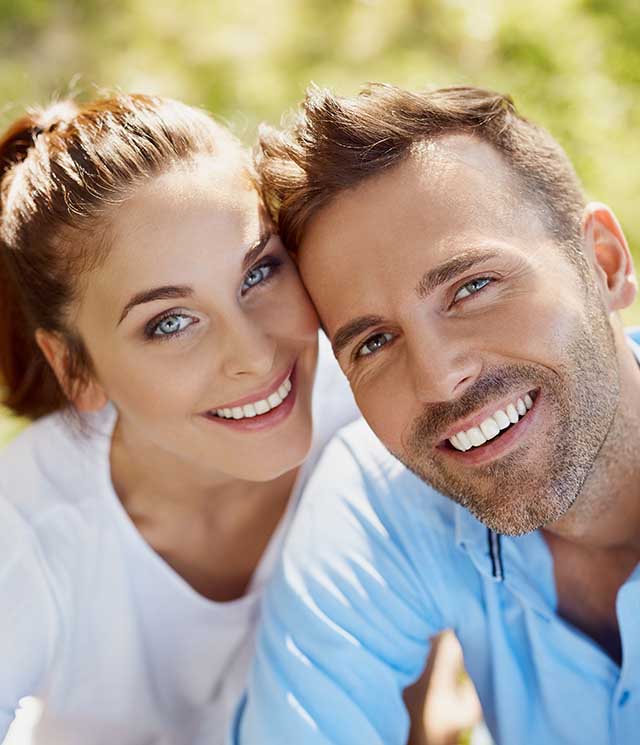 A young couple posing for a photo with smiles on their faces, set against a blurred outdoor background.