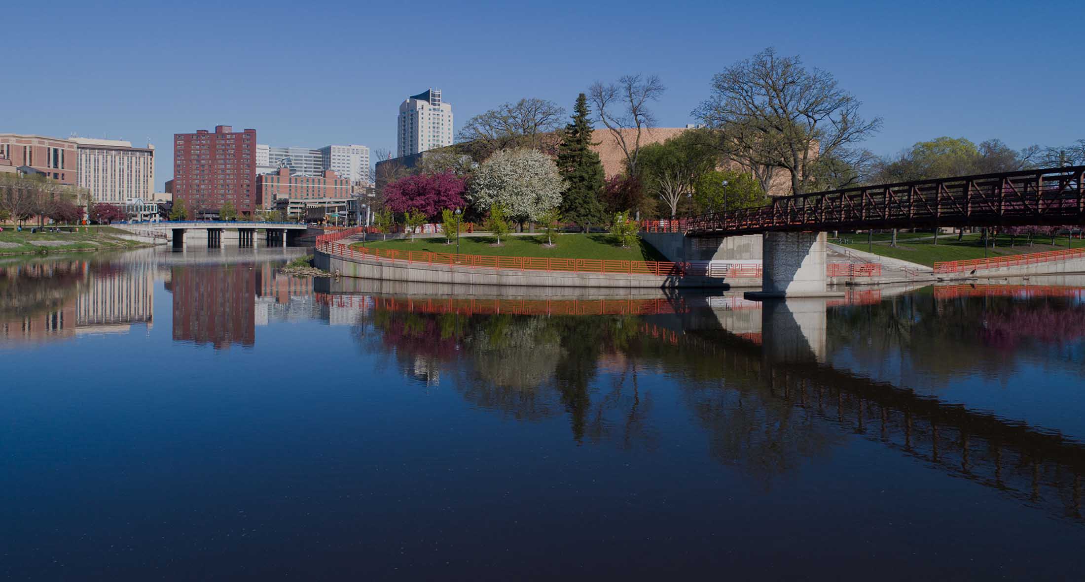 The image shows a scenic view of a river with a bridge crossing over it, reflecting the surrounding buildings and trees under a clear blue sky.