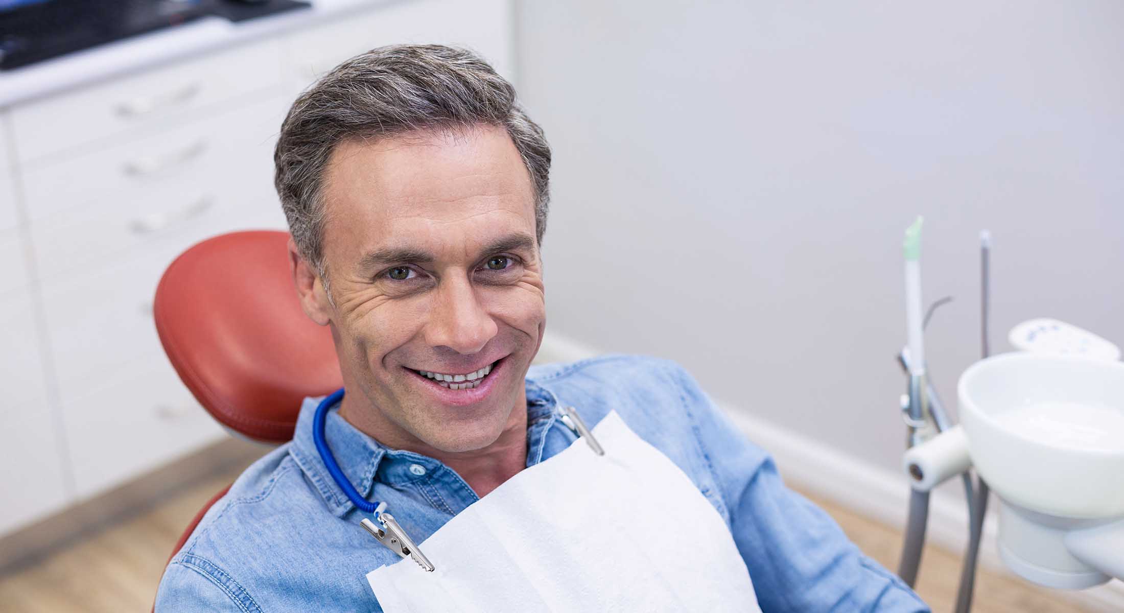 The image shows a man sitting in a dental chair with a smile on his face, wearing a blue shirt and blue jeans, while a dentist with a beard sits behind him, smiling and looking at the camera.