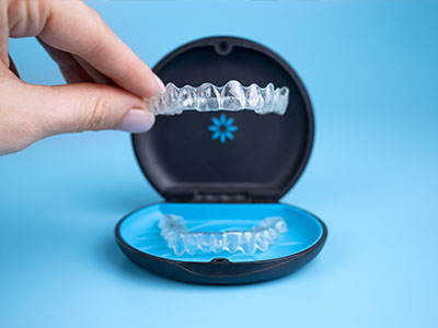 The image displays a hand holding an open case containing clear dental aligners next to a blue container with a transparent lid, which appears to be a dental appliance, possibly a mouthguard or a retainer.