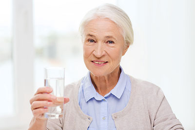 The image depicts an elderly woman holding a glass of water up to her mouth, smiling gently at the camera.