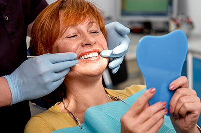 A smiling woman sits in a dental chair while a dental professional adjusts her teeth, with a blue mouthguard held up for inspection.