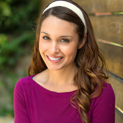 The image shows a woman with long hair smiling at the camera  she wears a purple top and has her hair styled down.