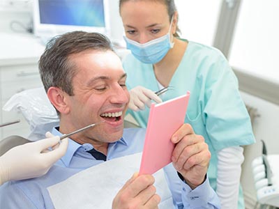 The image depicts a man sitting in a dental chair with a smile on his face, holding a pink card or booklet, while a female dental professional stands behind him, looking at him with a slight smile. They are both in a dental office setting, and the man appears to be engaged in an activity related to the card he is holding.