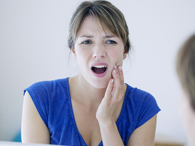 A woman with her mouth open, showing teeth, looking at her reflection in a mirror while holding her face with one hand.