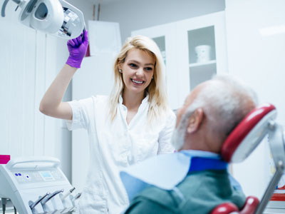 The image shows a female dental hygienist assisting an elderly man in a dental chair, with her hand holding up a dental mirror.