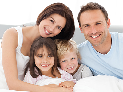 A family of four posing on a bed with smiles, featuring a man, woman, and two children.