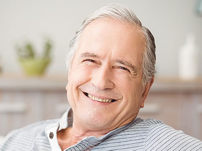 The image depicts an elderly man with gray hair, smiling broadly while sitting comfortably indoors.