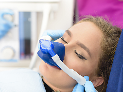 A woman receiving medical treatment with an oxygen mask on her face, attended by a healthcare professional wearing gloves and a surgical mask.