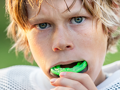 The image shows a young boy with blonde hair, wearing a football jersey, holding a green object near his mouth, possibly a sports-related item.